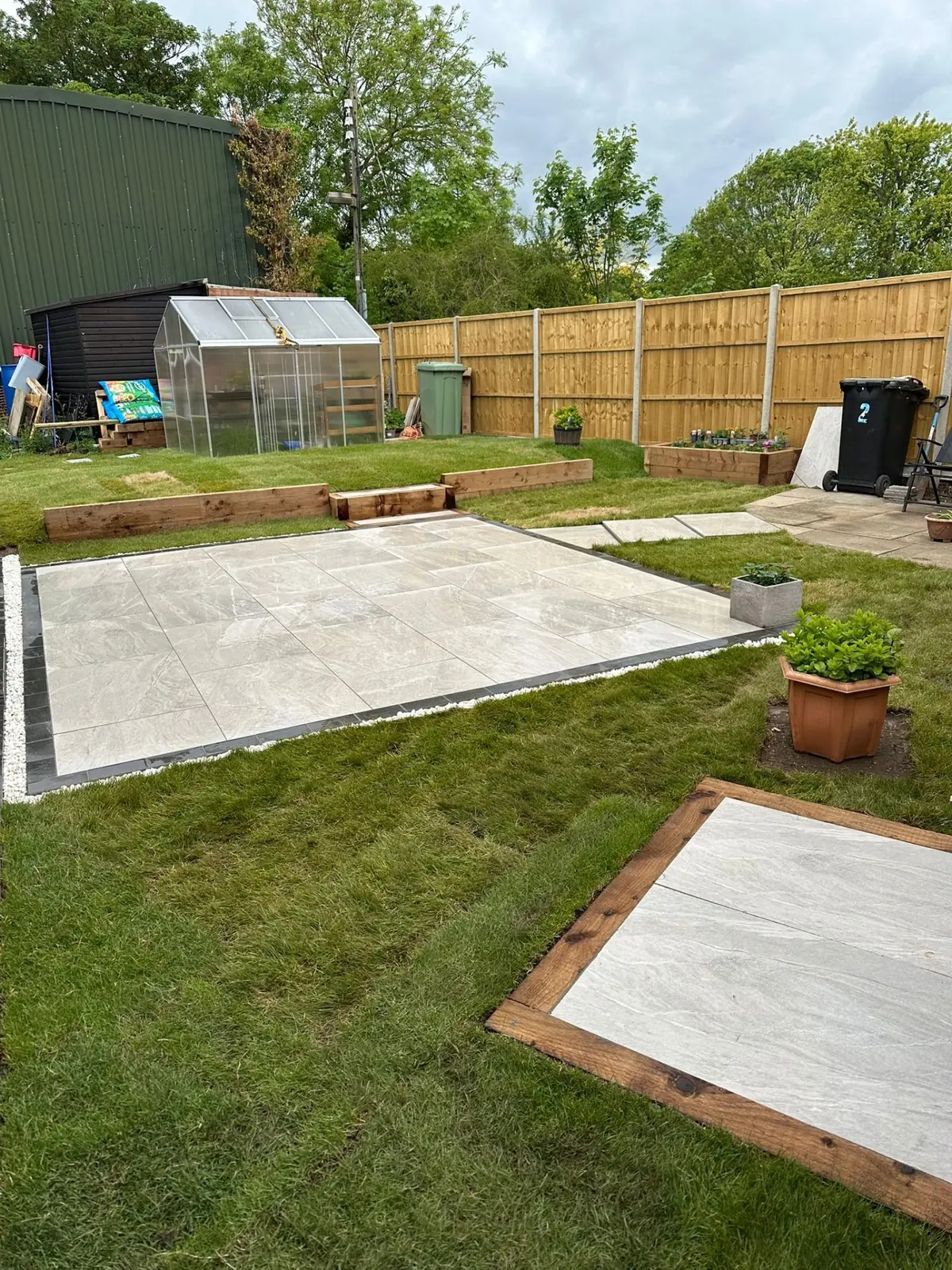 Grey porcelain patio with stepping stones, sleeper-edged lawn beds and greenhouse beyond