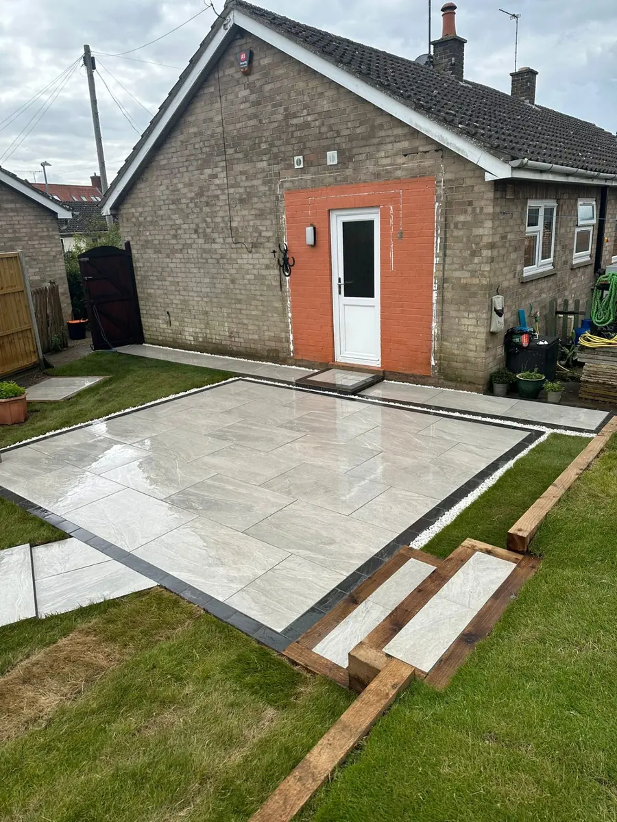 Grey porcelain patio with charcoal block-paving border, white pebble margin and timber sleeper steps down to the lawn
